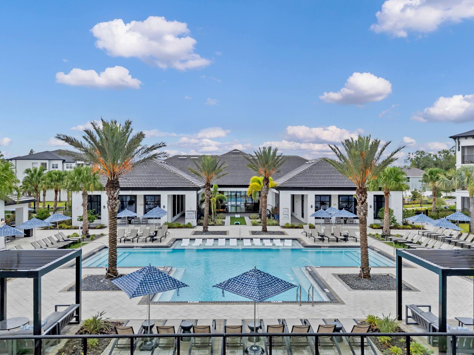Resort-style pool with lounge chairs, palm trees, cabanas, and blue sky with scattered clouds.