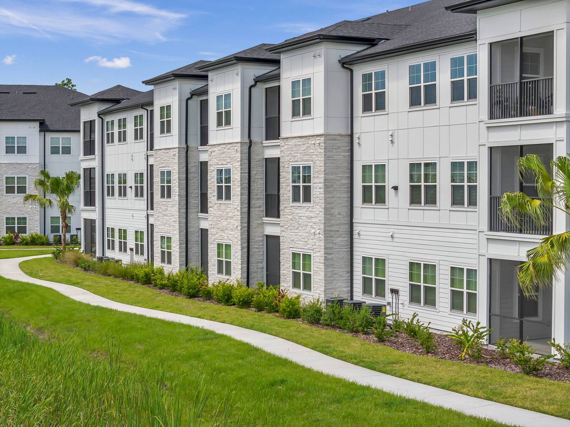 Modern three-story apartment building with white siding and stone accents, next to a green lawn and walkway.